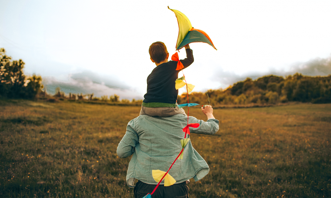 Father with his young son on his shoulders. The son is holding a kite as they run through a field.